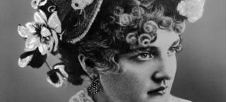 Image portrait of Baby Doe Tabor from the shoulders up. She is wearing a white lacy blouse with a flower attached on the right side of the high collar. She is wearing a very fancy hat on her head, that has white flowers on the right side of it and a large white ostrich feather on top. Her curly hair is seen beneath the hat, and she wears dangling earrings made of many small pearly beads.