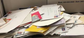 Photo of large stacks of letters and postcards addressed to Governor Jared Polis. There are additional boxes next to the mail, labeled "Box 2 Postcards." Visible postmarks and return addresses show that the correspondence has come from all around the country, including Providence, Rhode Island, West Hempstead, New York, and Pennsylvania.