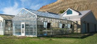 Photo of two greenhouse buildings made completely of framework and glass panels. The are connected in a perpendicular way to a primary one-story building. The greenhouses have 3 window ventilation fans on each side, and there is an entrance door at the short end of the greenhouse building. The property around the buildings is covered in green grass, and behind the buildings a brown bluff is visible and striking next to the bright blue sky.