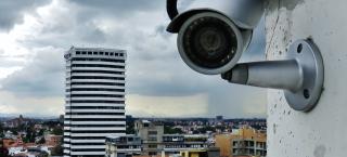 Photo of a security camera attached to the exterior wall of a high-rise building in a city. The security camera is looking directly at the viewer of this image.
