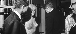 Photo of Robert Kennedy on the left of the image, standing next to Marilyn Monroe who is wearing the iconic nude-looking dress that is covered in rhinestones. President Kennedy stands next to her with his back to the camera and they are in a room with shelves filled with books. There is a partial image of a man captured on the right edge of the image. They are talking and smiling.