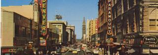 Image of a picture postcard of Downtown Denver around 1950-1960. The photograph has been taken from an intersection along Sixteenth Street. Signs on buildings read "Paramount (Theatre)" "Denver" "Skagg's" and "Swift's Ice Cream," among others. Cars are travelling in one direction along Sixteenth, toward the D&F Tower in the distance.