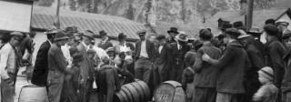 A group of men in period 1920s clothing stand around a wooden barrel of alcohol while it is poured into the gutter.