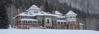 A turn of the century manor with a broad, snow-covered lawn and a dense forest behind it.
