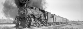 Black and white photograph of steam engine billowing black smoke and hauling a train of cars.