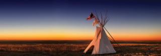 A tipi at sunset. Over the tipi fly two flags: the United States flag, and the white flag of peace.