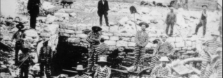 Black and white photo of men in striped prison uniforms digging a large hole. Two guards in dark uniforms observe. A river and mountains are in the background.