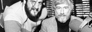 Two bearded men pose with a microphone in front of them. The man on the left has had an X marked over his face on the print.