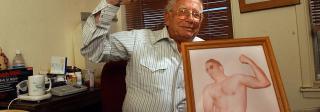 Dr. Stanley Biber flexing his right arm in his office. In the foreground is a framed photo of him as a younger man also flexing his biceps