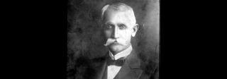Black and white studio portrait of a man in a suit with a mustache looking into the camera
