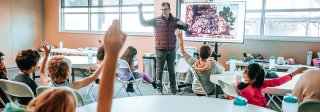 A class of students look attentively at a male teacher facing camera. He's pointing to a photo of petroglyphs while many children raise their hands.