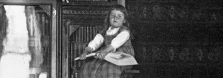 A young girl (Josephine Evans) sitting on a ladder with a book on her lap. Behind her is a filled bookshelf.