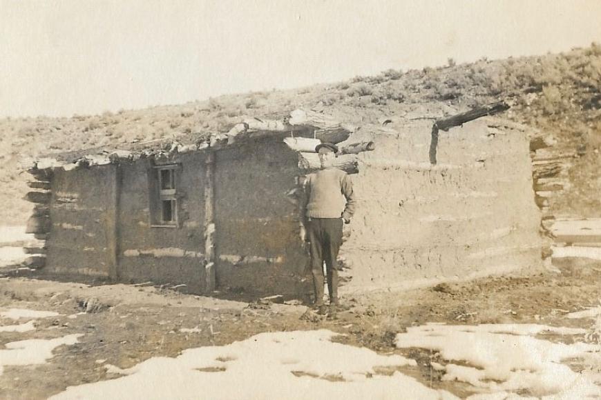 Historic image of man standing in front of a log and adobe cabin in the snow.