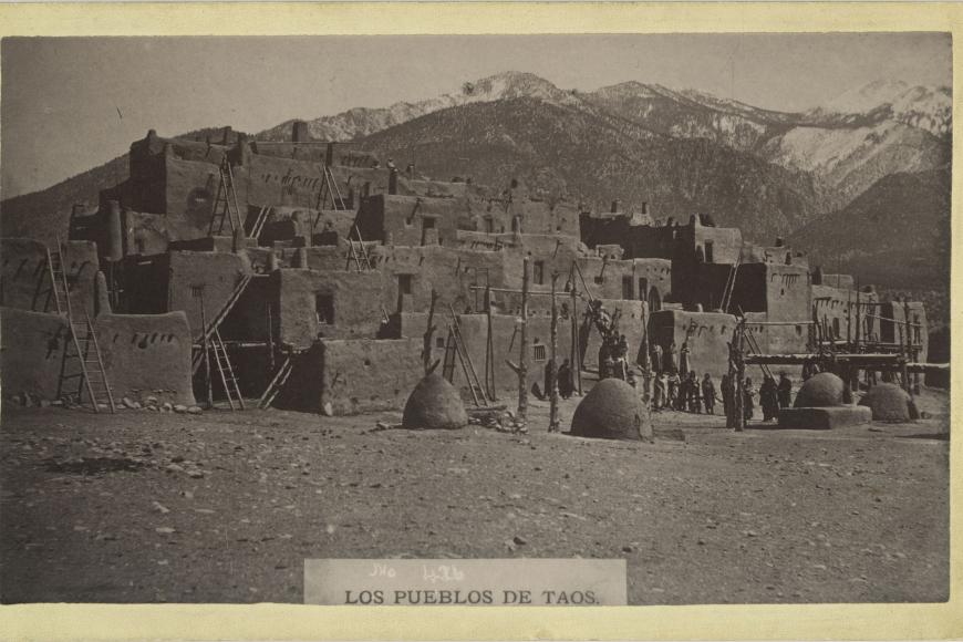 Photograph depicting Taos American Indians standing next to large adobe structures, photographed in Taos Pueblo in New Mexico by an unidentified photographer, circa 1900-1920. The group is mostly wearing blankets. Some stand on the ground, while others pose on the ladders leading to the adobe structures. Beehive ovens, also known as Hornos, are in the foreground.