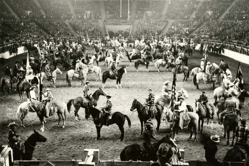 Photo of the indoor arena at the National Western Stock Show, full of dozens of riders in cowboy apparel on horseback, celebrating the inaugural event. The stands are packed with spectators.