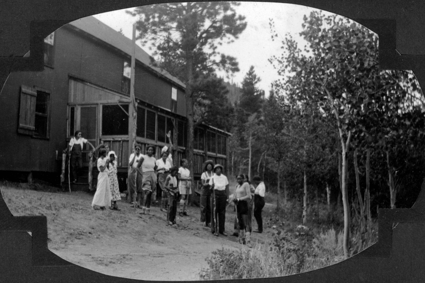 A group of African American teenage campers and their counselors stand near a camp building at Camp Nizhoni in Lincoln Hills (Gilpin County), Colorado. Some hold large sticks. They wear shorts, overalls, pants, shirts, dresses and hats.