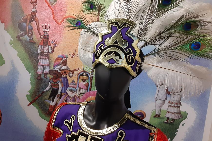 A male mannequin wears a bright colored vest or tunic covered with sequins. On its head is a feathered headress with traditional mesoamerican patterns. Behind it, a wall mural depicts dancers of various traditions on a map of Mexico.