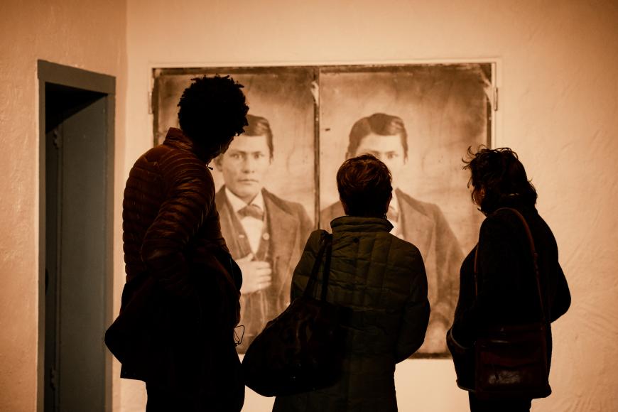 Three people observe a projected image of an antique photograph, depicting a seated Native American man in western dress. 