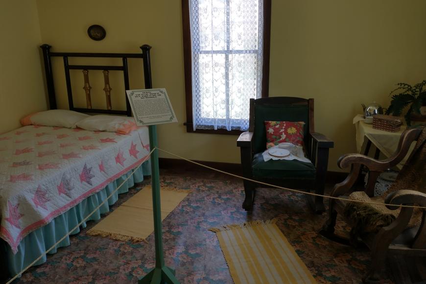A bedroom display at a house museum. There is a collection of antique furniture, including a bedframe, in a small room.