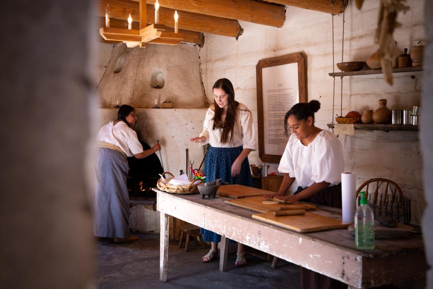 A small kitchen with adobe walls. Three women in traditional 1800s hispano dress are preparing a meal. This room is a recreation of an original kitchen.
