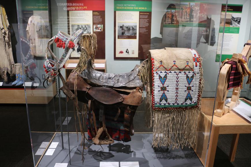 A saddle on display in a glass case. The saddle is of Native American (Ute) make, with fine beadwork decorating it. Behind the case containing the saddle are panels with further information.