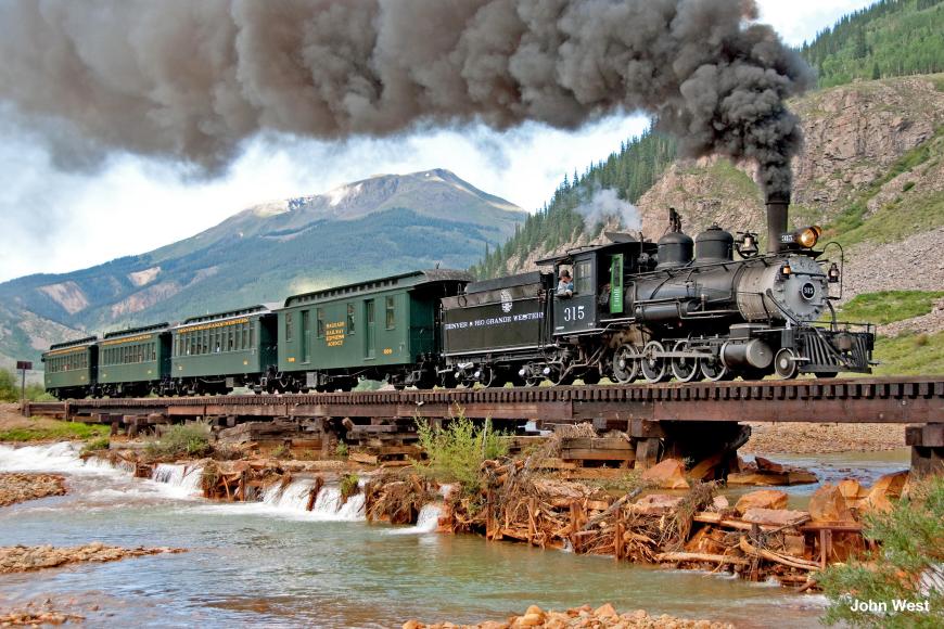 A color photo of steam locomotive Engine 315, after restoration. The engine is under steam near Durango and hauling coal wagons. Mountains are visible behind.