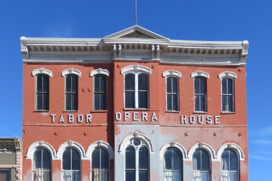 Facade of the Tabor Opera house in Leadville, CO. The three story building has shops in the bottom floor, and a large flagpole on the roof. It is constructed of red sandstone in a neoclassical style. On the facade are the words "TABOR OPERA HOUSE" in tall letters.