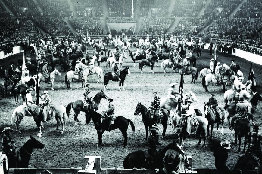 A large stadium arena surrounded by bleachers. On the floor of the arena, a multitude of horses and riders are gathered, most wearing "cowboy" clothes.