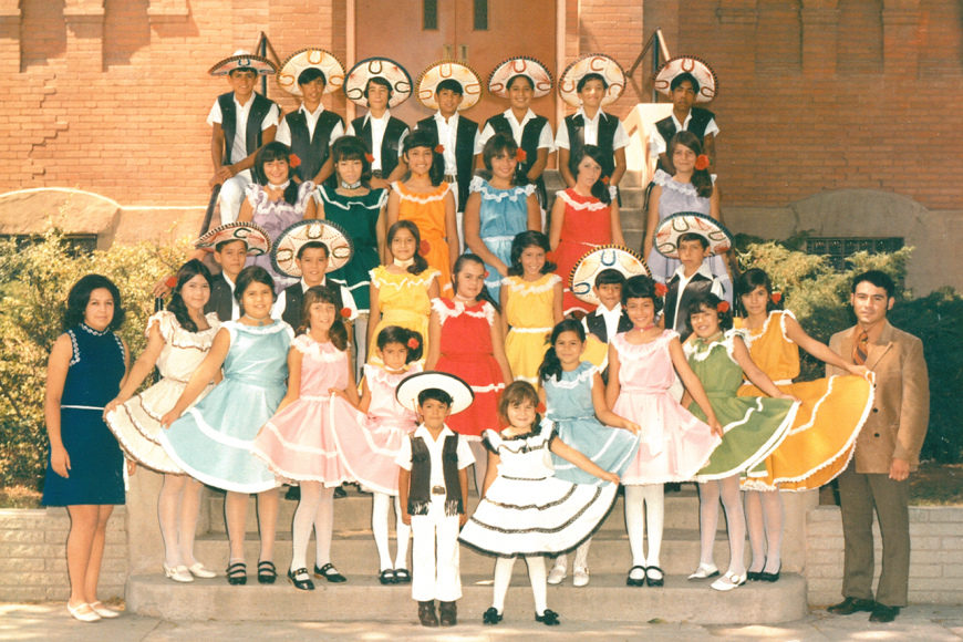 A Folklórico group from Pueblo poses on stairs before a building. The girls are dressed in traditional dresses of many colors. The young men wear black embroidered suits and white hats.