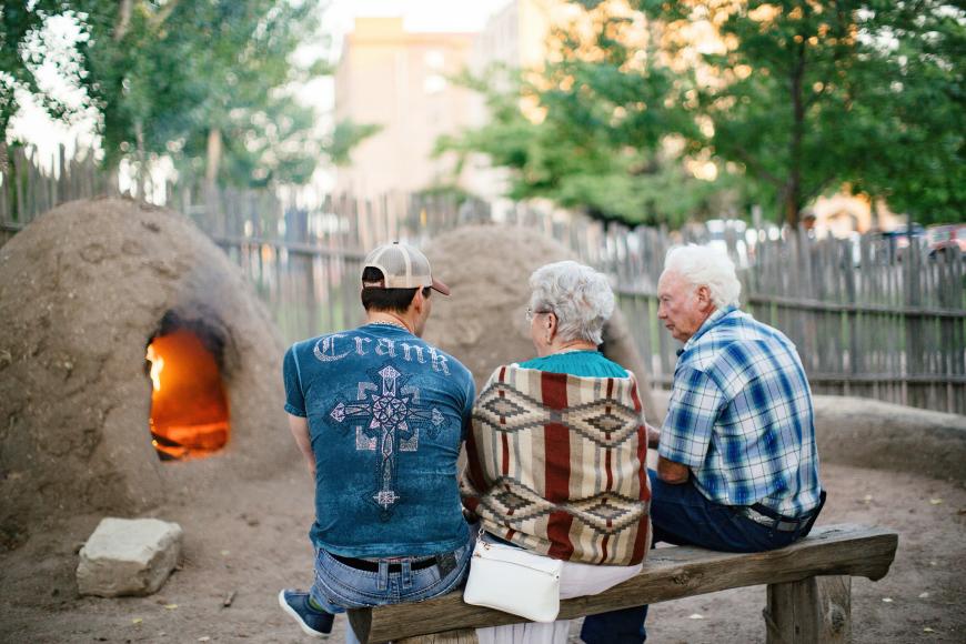Three figures are seated on a wooden bench in front of a lit horno- a type of beehive-shaped outdoor clay brick oven, made of adobe. There is a fire within the horno. In the background is a wooden coyote fence and several trees.