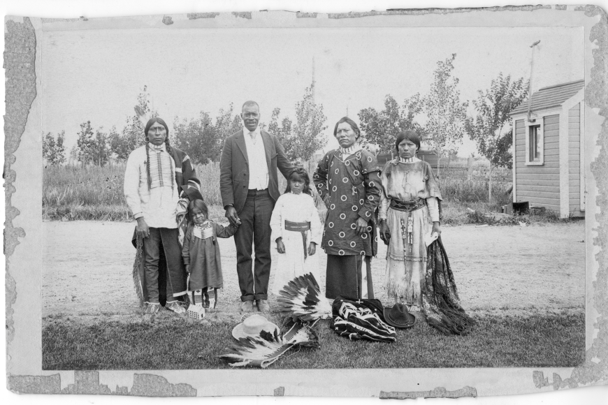 A group of people stand in front of a farm or small homestead. One is a black man in a business suit. Around him stand several Indigenous (Ute) adults, and near him stand two  Ute children.