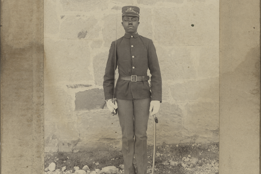 A Black soldier stands at attention in his parade uniform, with a saber in one hand.