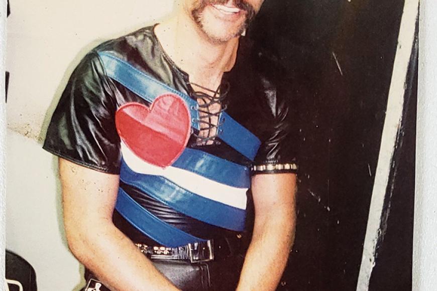 A man with a mustache wearing 1980s/90s leather scene fashion, including a leather shirt, hat, and gloves.