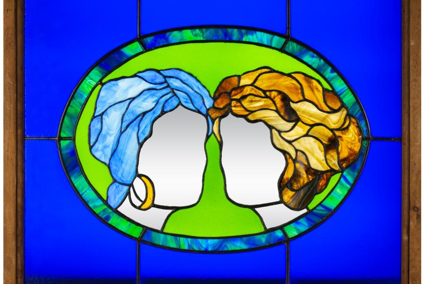 A stained glass window, depicting two women's heads facing each other. One has blue hair, the other has blonde hair. They are faceless.