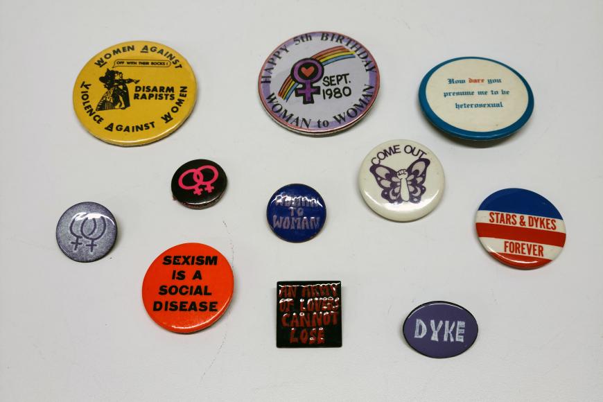 A collection of colorful political buttons on a white surface. They read: "Women Against Violence Against Women (Disarm Rapists)" ; "Happy 5th Birthday Woman to Woman Sept. 1980" ; "How dare you presume me to be heterosexual" ; "Woman to Woman" ; "Come Out" ; "Stars & Dykes Forever" ; "Sexism is a Social Disease"; "An Army of Lovers Cannot Lose" ; and "Dyke"
