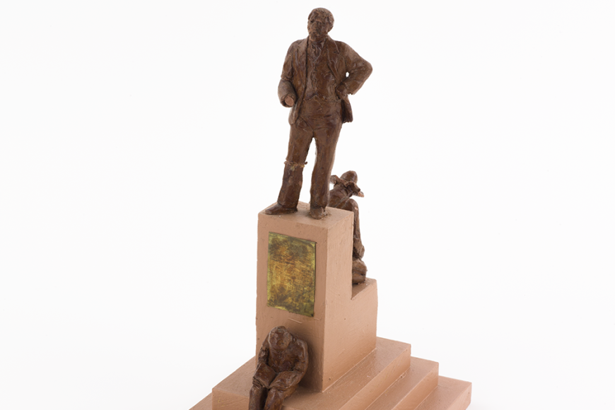 A statuette depicts a man in three stages. The lowest level depicts him crouched in bondage, wearing the chains of a slave. The second level depicts him crouched with a gold pan. The third level at the peak of the plinth shows him standing tall and proud.