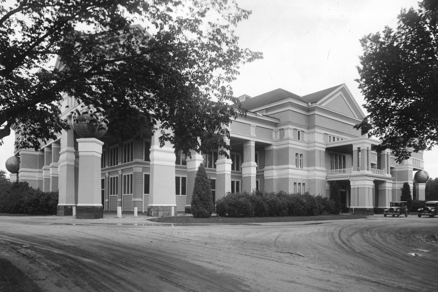 A black and white photo of a large building with geometric features. Its exterior is dominated by pillars and its peaked roof is higher than the trees around it. There is a dirt driveway leading up to the building where a car is parked.