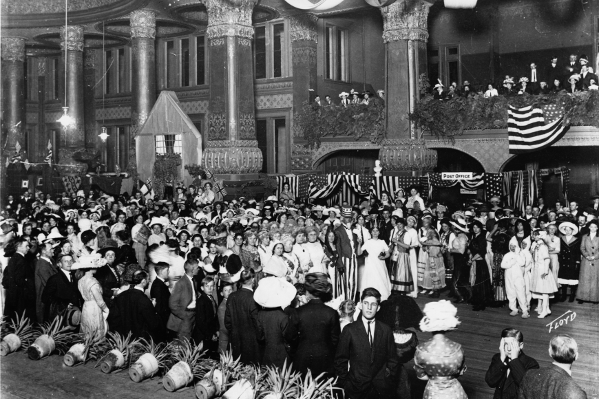 A celebration being held inside the Mineral Palace. A large crowd fills the open atrium between the pillars. Near the center of the crowd are individuals dressed as Uncle Sam, Columbia, and Lady Liberty.