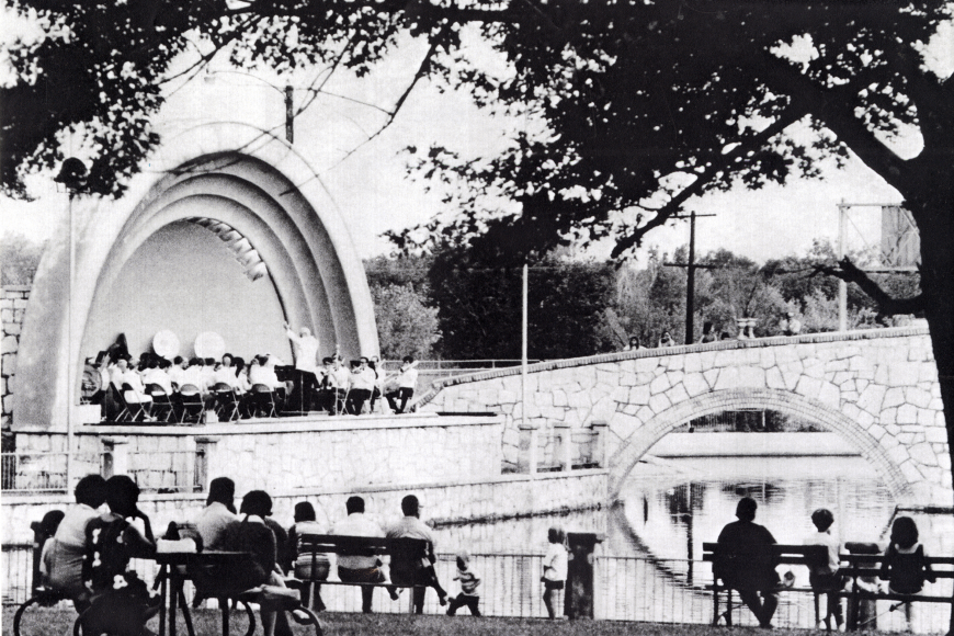 A performance in the Mineral Palace Bandshell, likely by the city's municipal band. People are seated on benches across the water, watching.