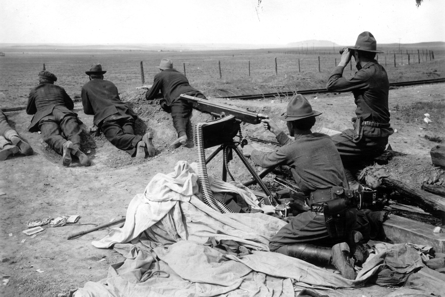 A man in Colorado National Guard uniform a machine gun nest. He is pointing it away from the camera. Another National Guardsman kneels next to him with binoculars. Three more are laying prone in front of the machine gun.
