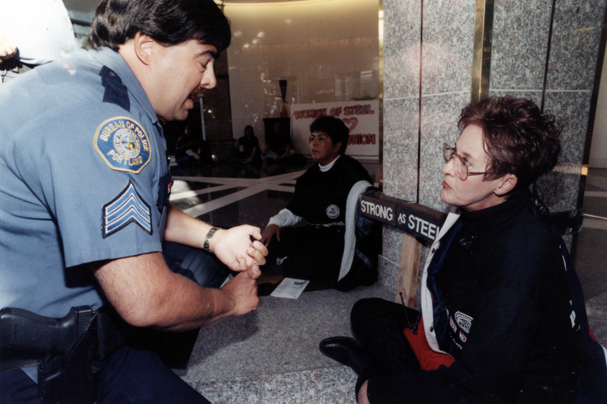 A protestor speaks angrily to an irate police officer. The protestor is holding a sign which reads "Strong as Steel."