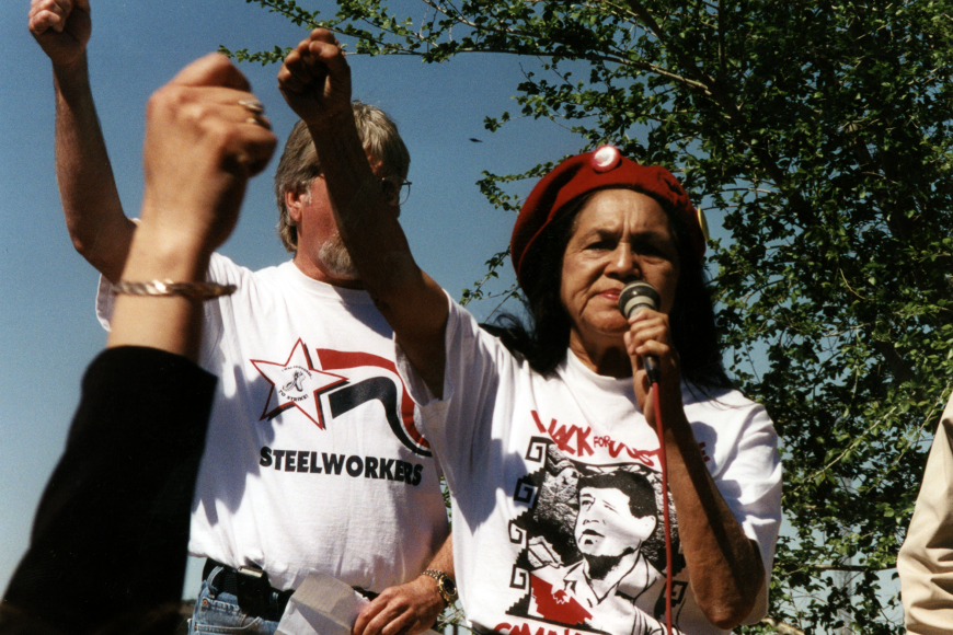 Two protestors raise their fist. The woman in front is holding a microphone and wearing a beret. Their shirts read "Walk for Justice" and "Steelworkers."