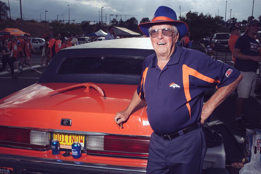 A Broncos superfan wearing a jersey standing next to his orange classic car.