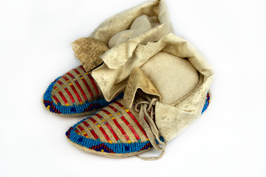 A pair of children's shoes made with animal hide and beaded with blue, red, and yellow beads in a striped pattern.