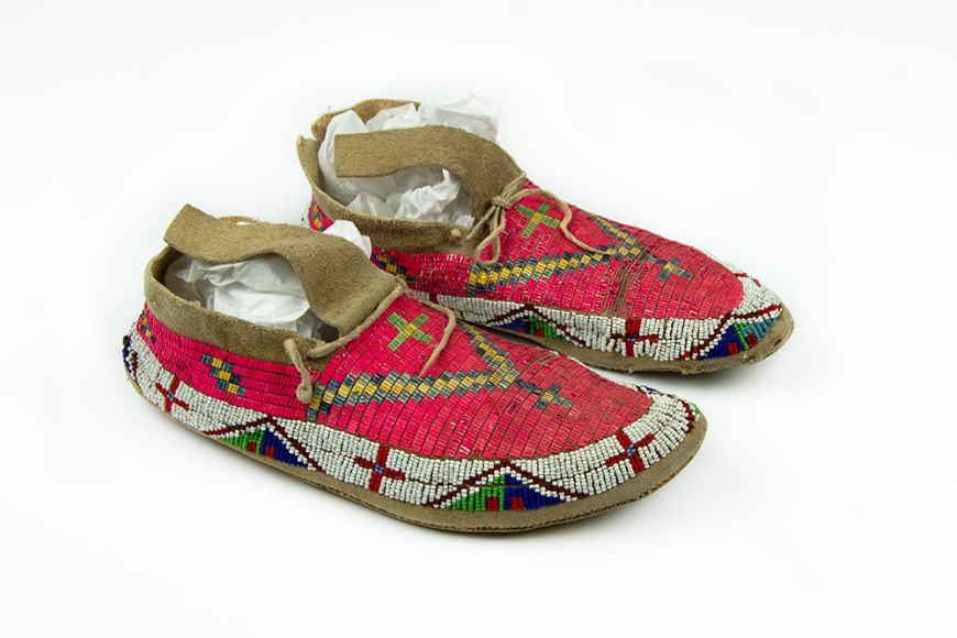 A pair of red children's shores made from animal hide. It is decorated with cross and triangle patterns with red, blue, green, and white beads.