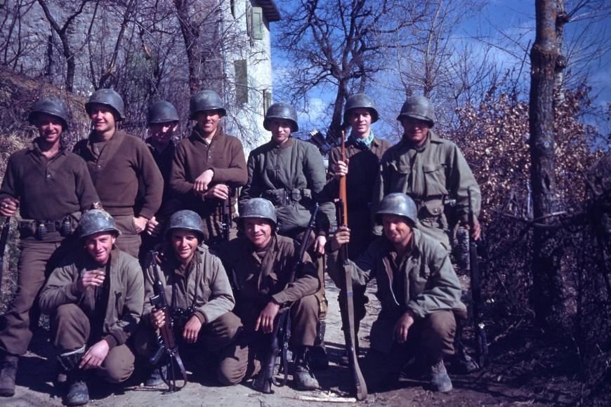 A group of 11 soldiers posing for the camera at camp hale and smiling. They have guns and bows and arrows, as well as grenades.