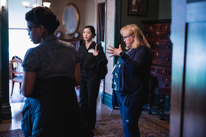 A small group listens to a female tour guide as she speaks at Center for Colorado Womens History. She has a dark wooden background behind her and is motioning at something out of frame.