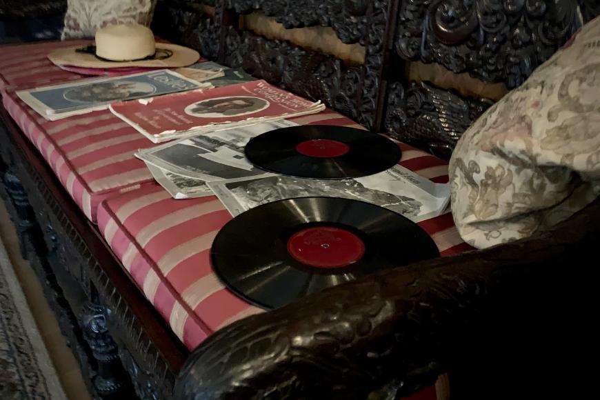 A dark wooden bench carved ornately. On it sits multiple vinyl records and record slips. The cushion is red and white striped.