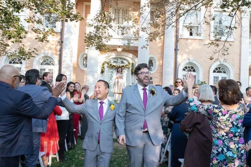2 grooms on their wedding day in grey and pink suits. They walk through a crowd of celebrating attendees from the Grant Humphrey's Mansion lawn.
