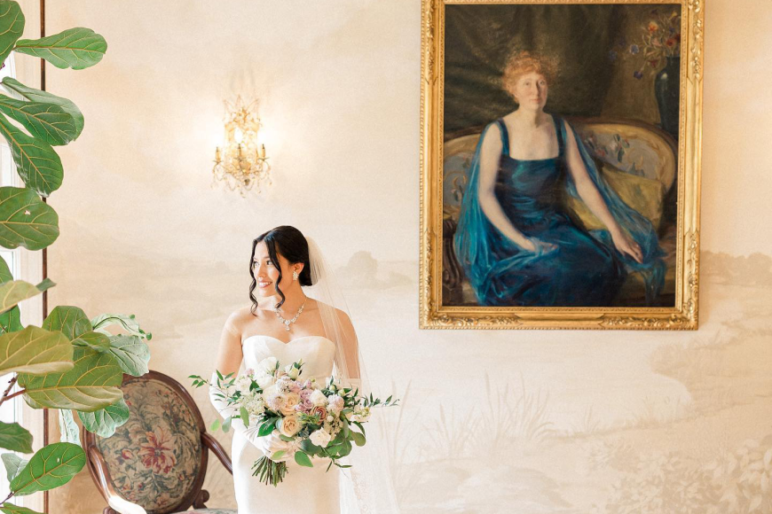 A bride inside Grant Humphreys mansion in her dress and holding her bouquet. She stands by a plant and old portrait of a woman.